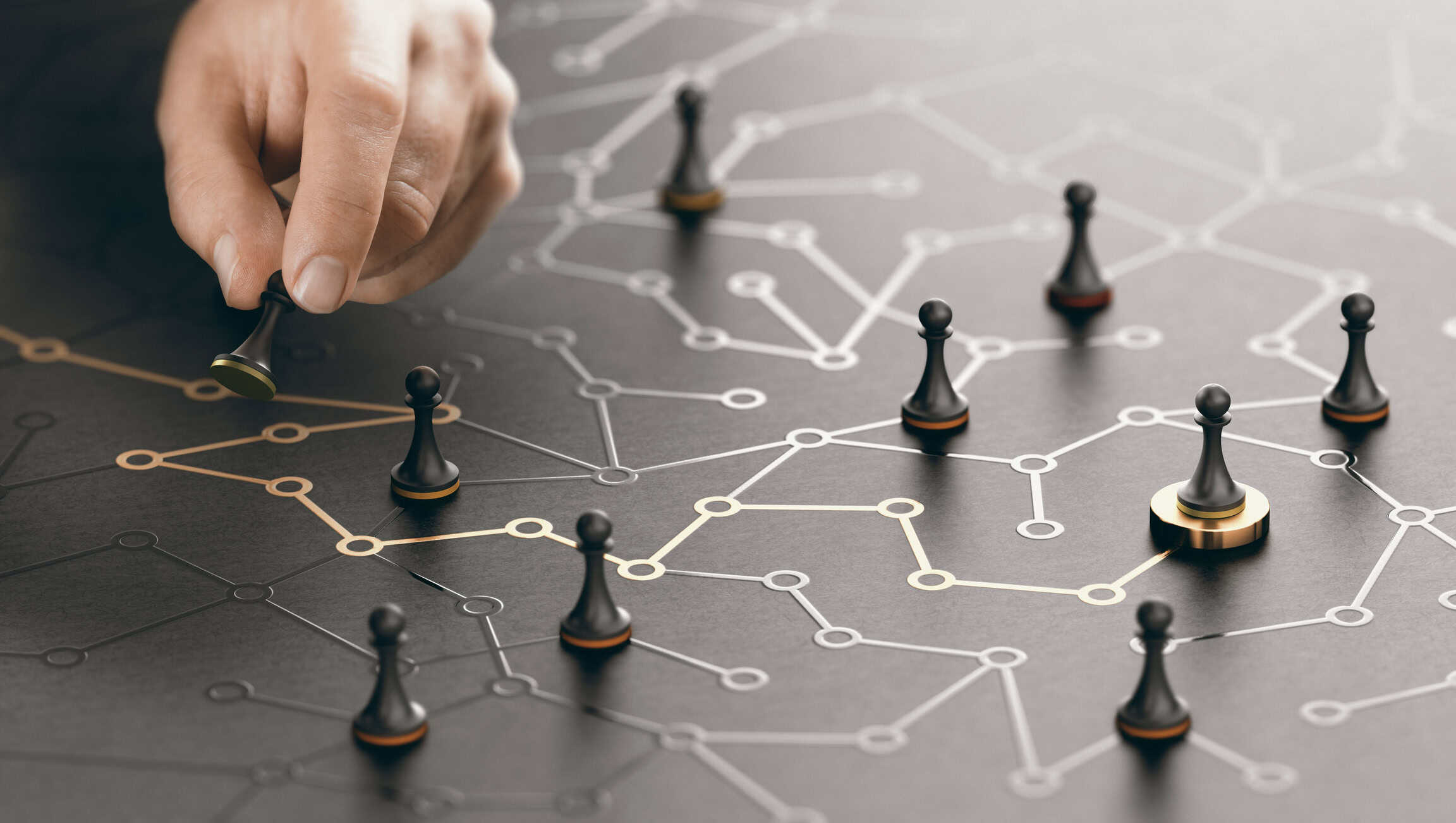 A hand moves a black chess pawn with a gold base on a dark, textured surface featuring a network grid of interconnected white and gold lines and circles. Other black pawns are scattered across the grid.