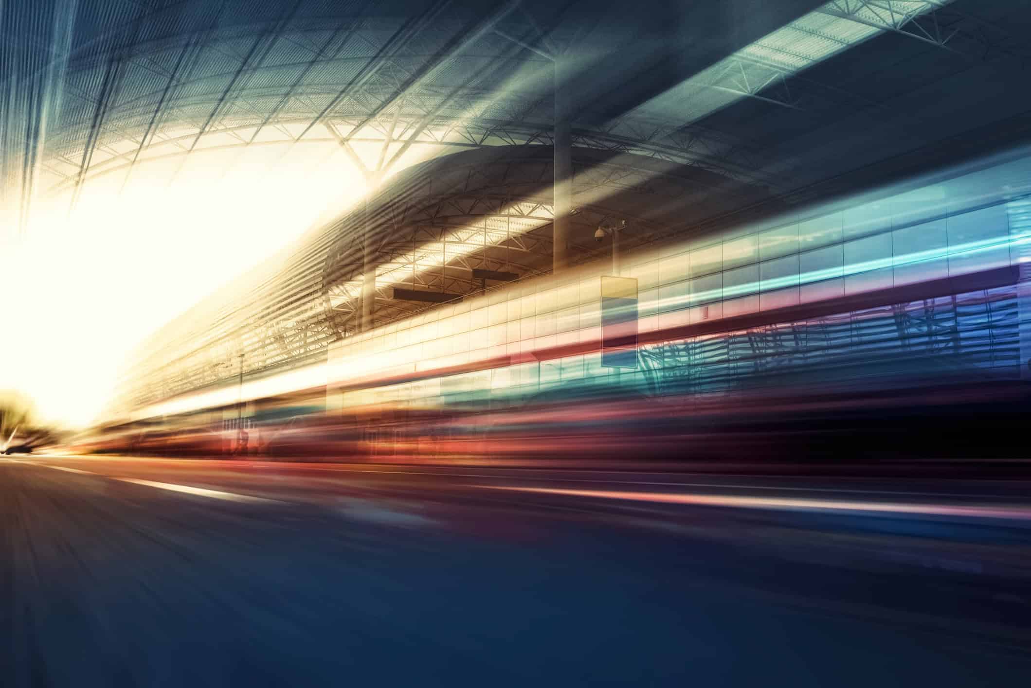 An abstract image showing motion blur, with bright light on the left, streaking colors across the center, and a blurred architectural structure above, suggesting high speed.
