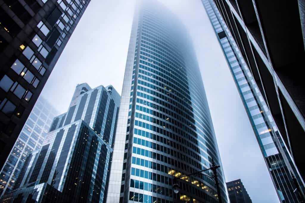 Low-angle view of multiple modern glass skyscrapers, with the tallest central building disappearing into dense fog.
