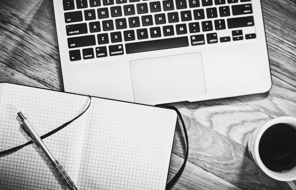 Black and white image of a laptop, an open grid notebook with a pen, and a cup of coffee on a wooden desk.