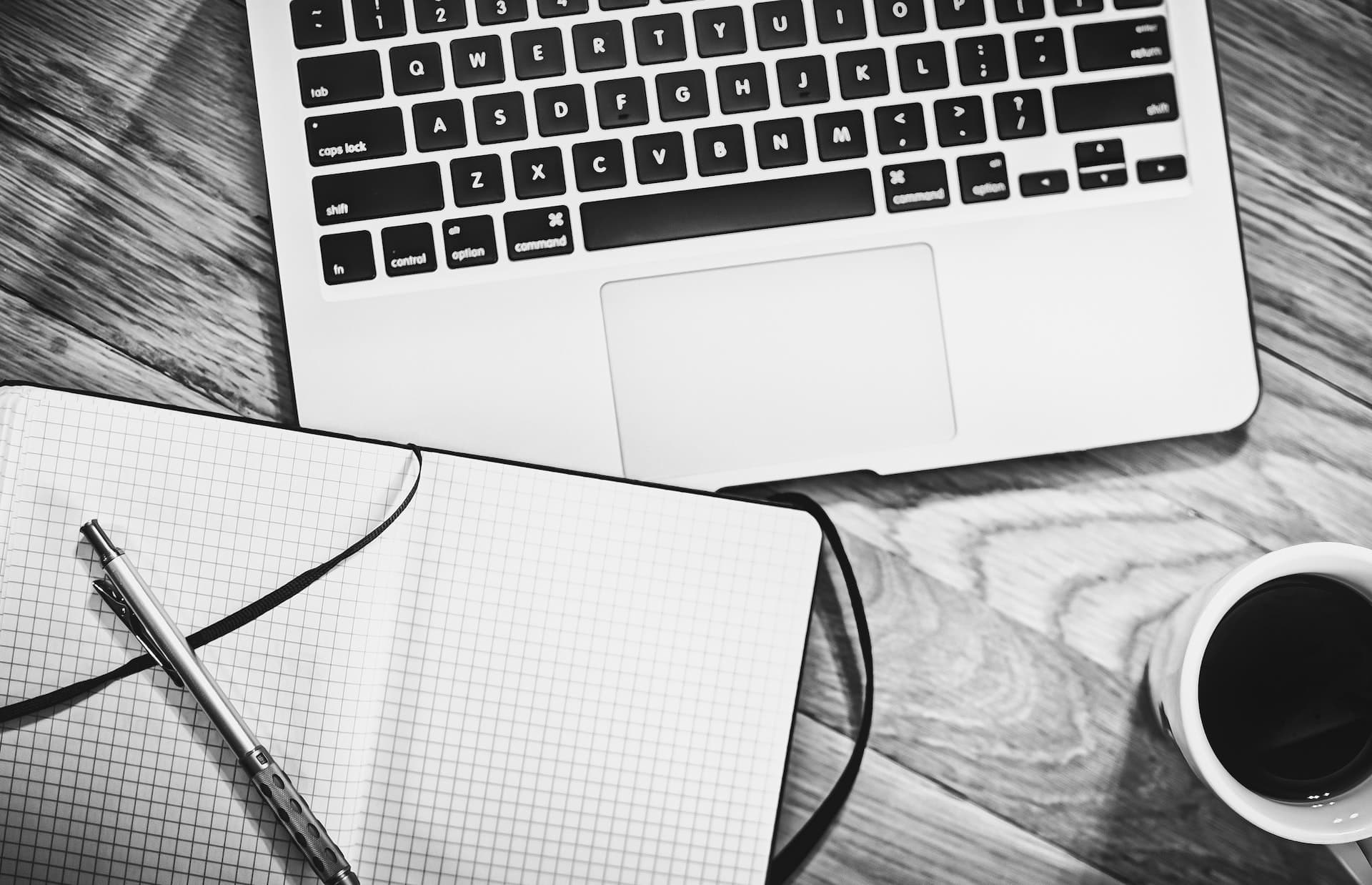 Black and white image of a laptop, an open grid notebook with a pen, and a cup of coffee on a wooden desk.