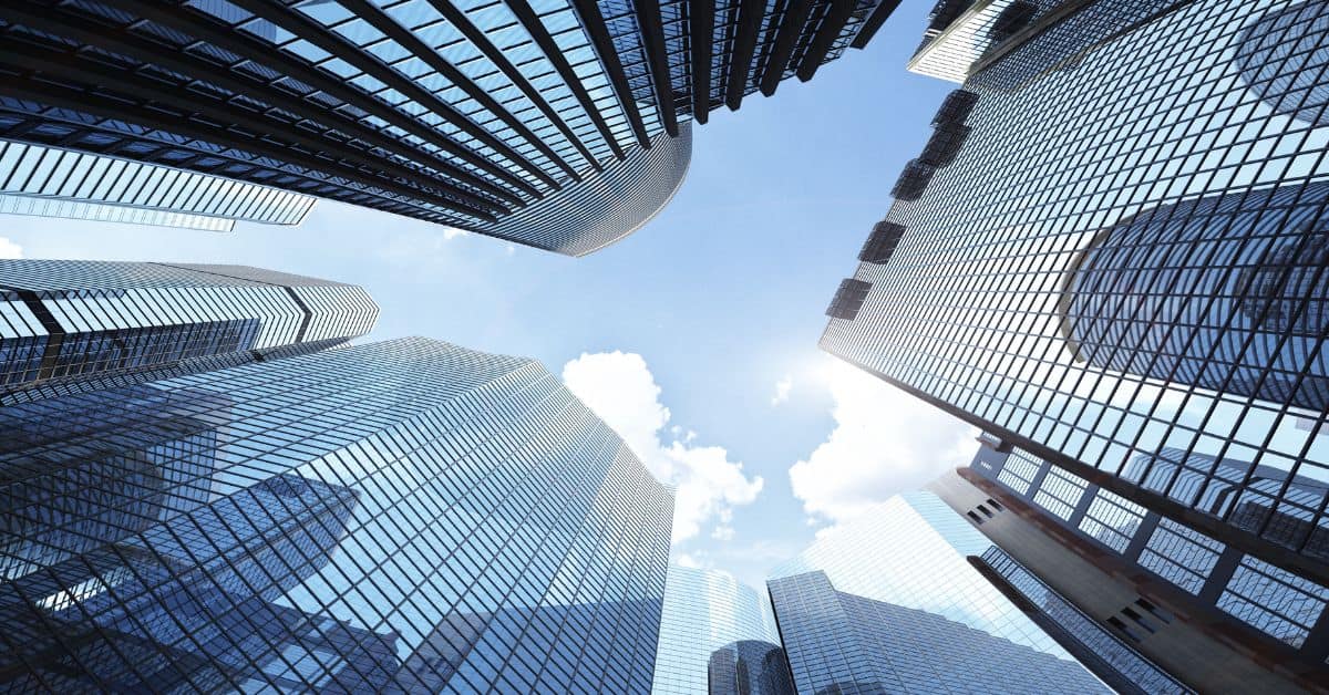 Low-angle view of multiple glass and steel skyscrapers against a bright blue sky with a visible sun flare.