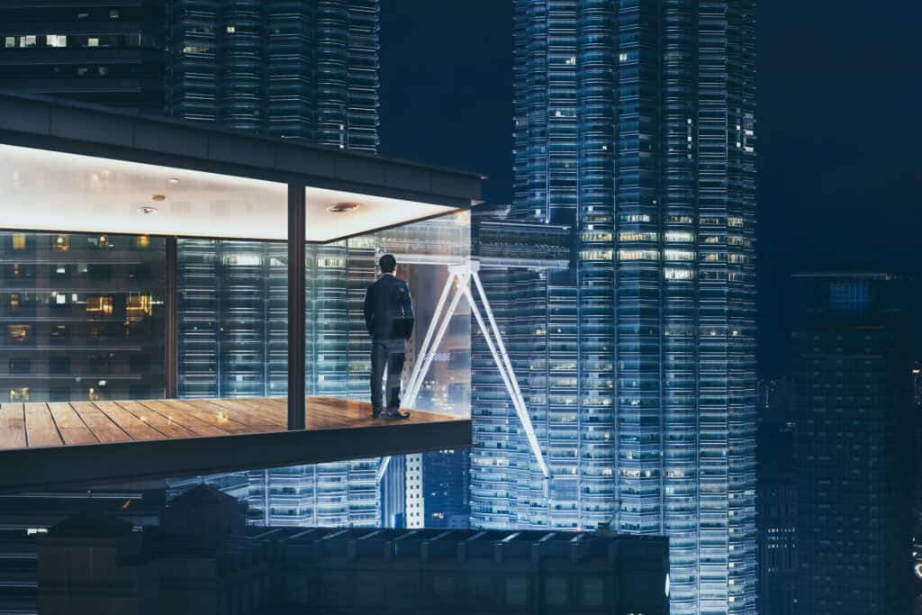 Rear view of a man in a suit standing on a glass balcony, looking at two brightly lit skyscrapers at night.