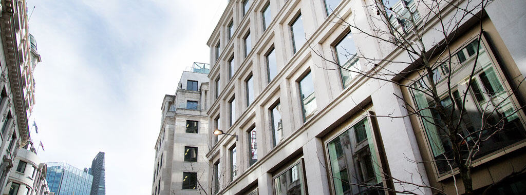 Upward view of light-colored buildings with rectangular windows and bare tree branches against an overcast sky. A modern glass skyscraper is visible in the background.