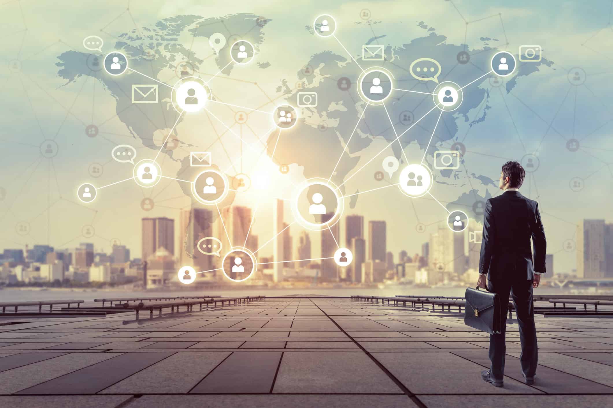 A businessman in a suit stands on a paved area, looking at a cityscape with a glowing world map and network of communication icons overlaid in the sky.