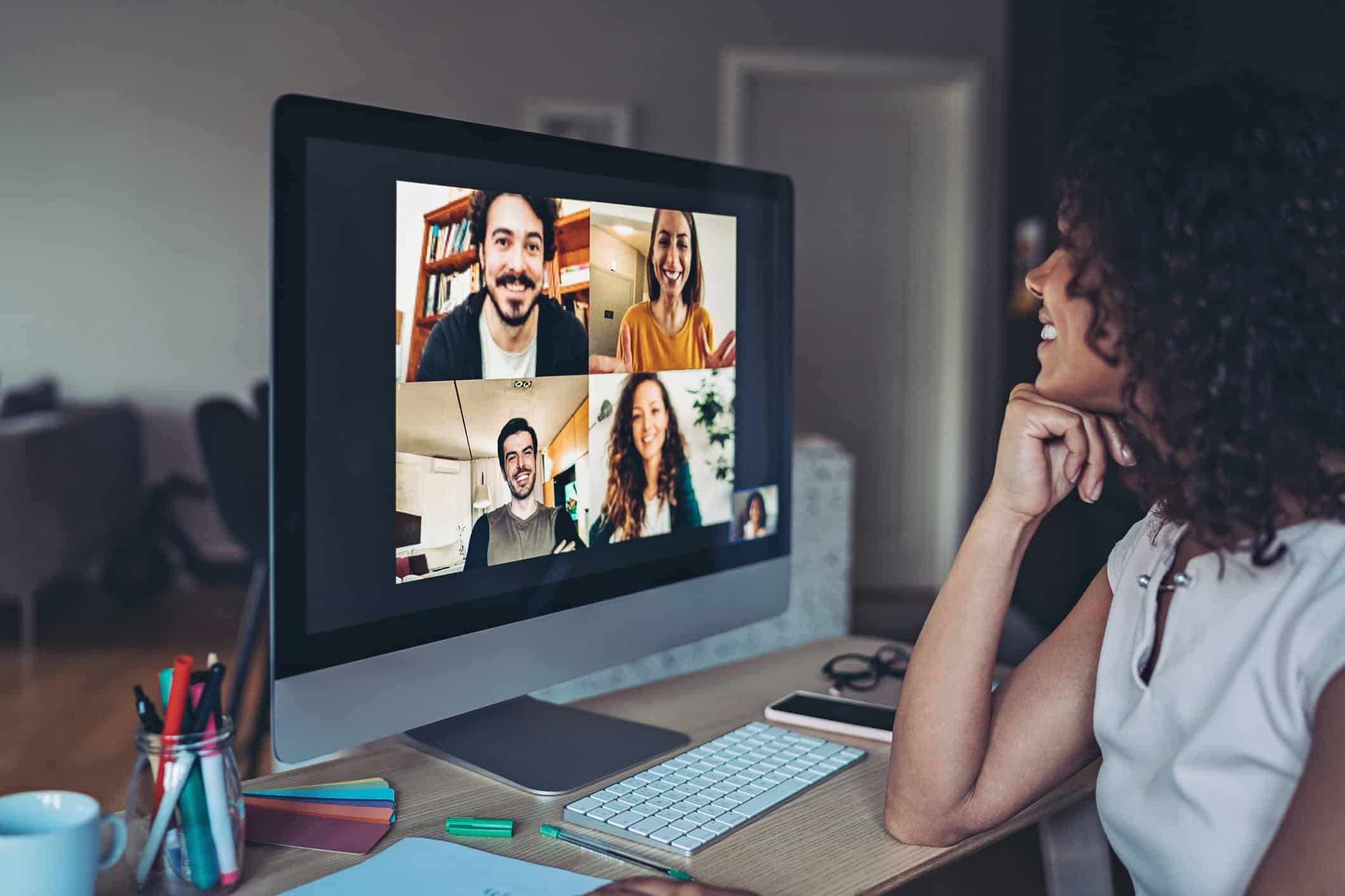 A smiling woman with curly hair participates in a video conference call on her desktop computer, showing multiple participants in a grid.