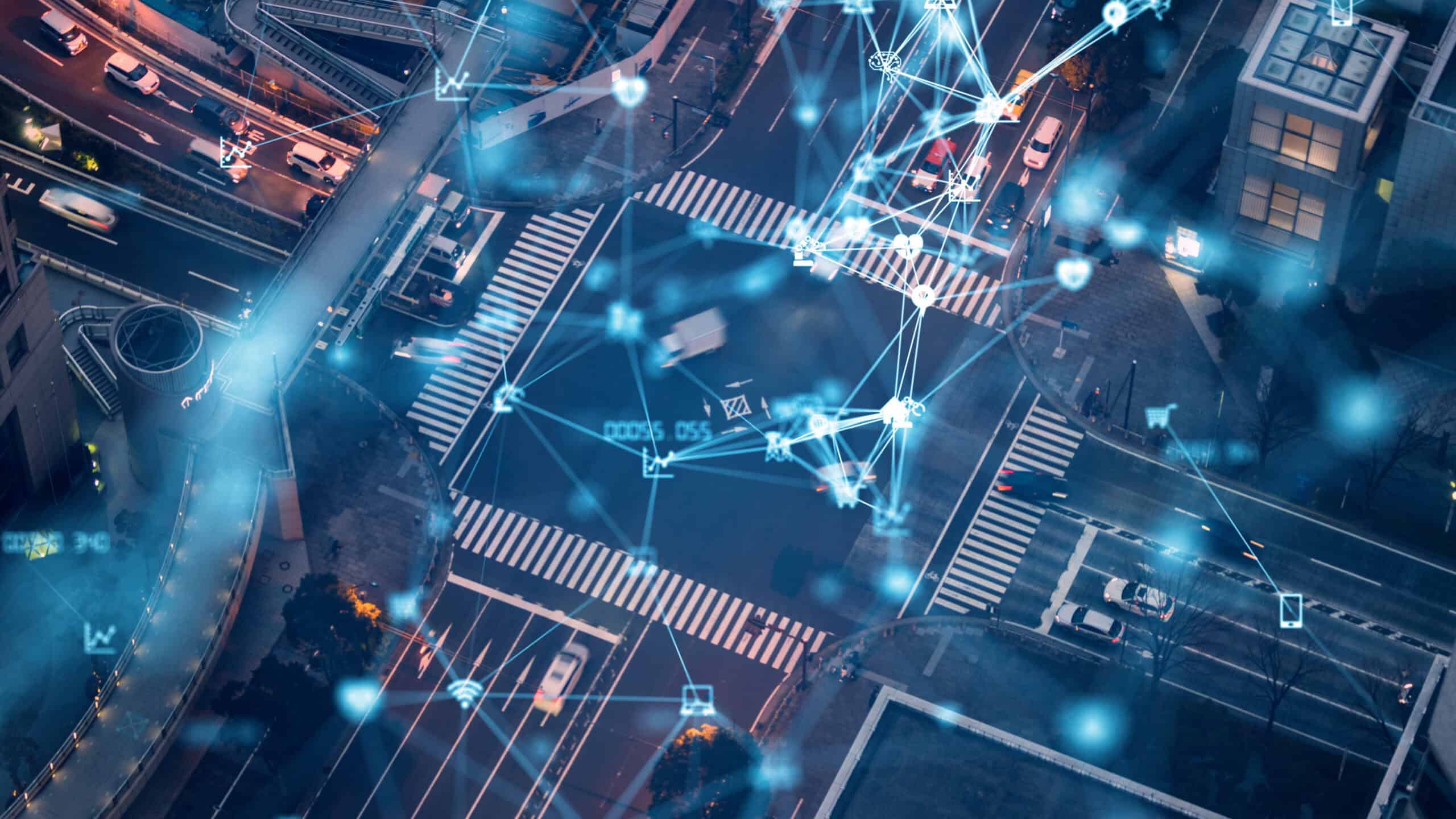 Aerial view of a city intersection at night with cars, overlaid with a glowing blue digital network of interconnected lines and various technology icons.