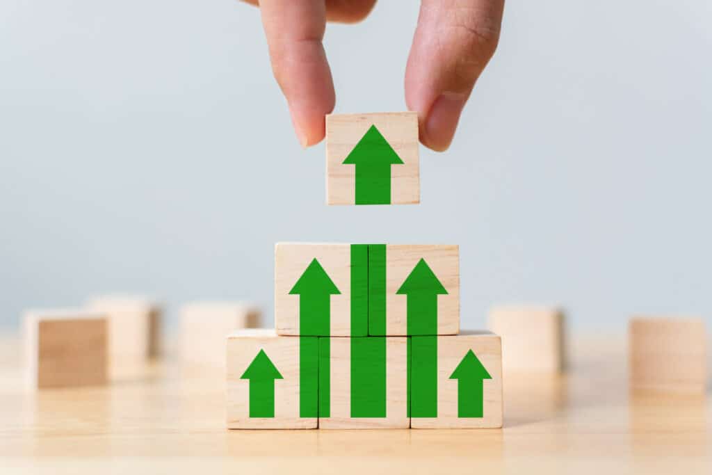 A hand places a wooden block with a green upward arrow on top of a stack of blocks, forming a rising bar graph.