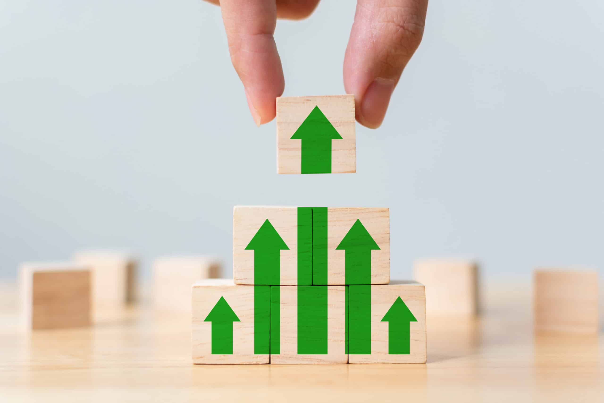 A hand places a wooden block with a green upward arrow on top of a stack of blocks, forming a rising bar graph.