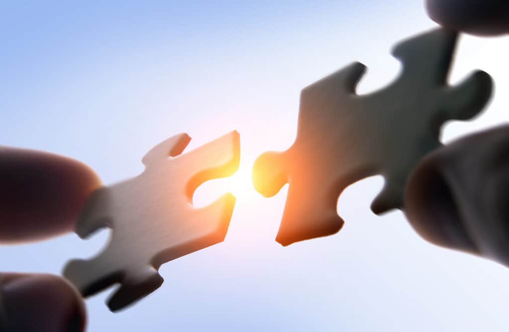 Two puzzle pieces held by fingers, about to connect, with a bright orange light emanating from the gap between them against a light blue sky.