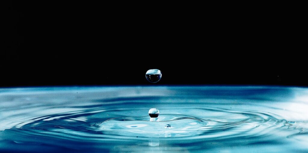 Two water drops, one suspended and one impacting a blue water surface, against a black background.