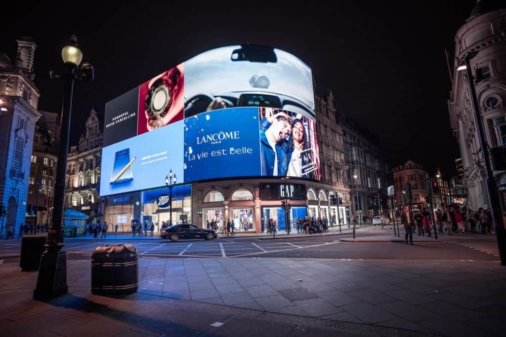 Night view of Piccadilly Circus in London, featuring large, brightly lit digital billboards on a curved building, with street-level shops, cars, and pedestrians below.