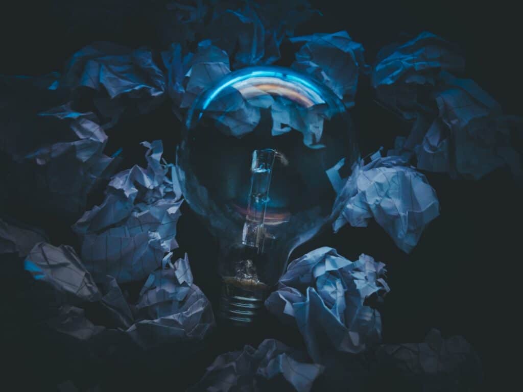 A clear light bulb, unlit, surrounded by crumpled white papers on a dark background, with blue reflections on the bulb.