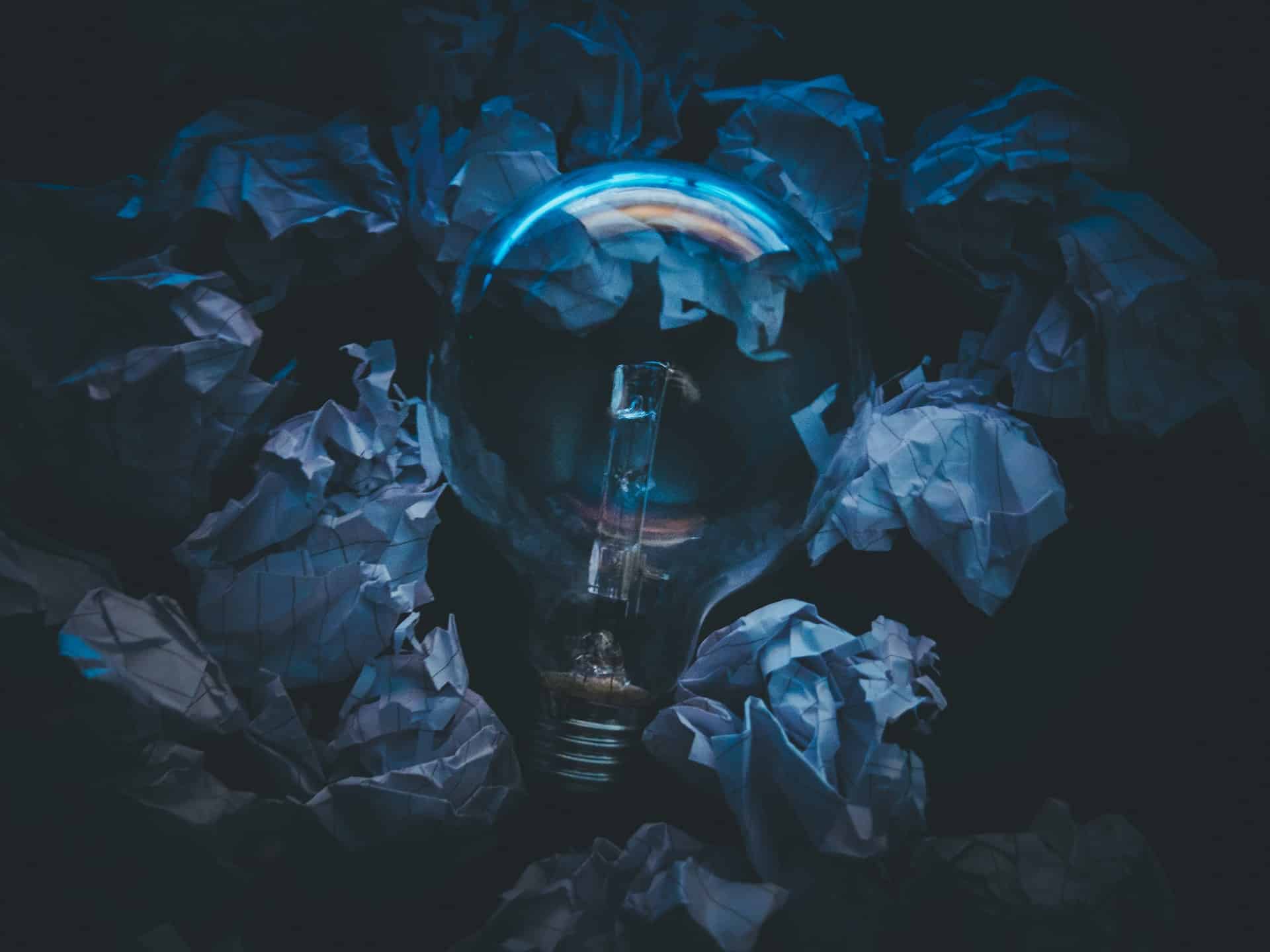 A clear light bulb, unlit, surrounded by crumpled white papers on a dark background, with blue reflections on the bulb.