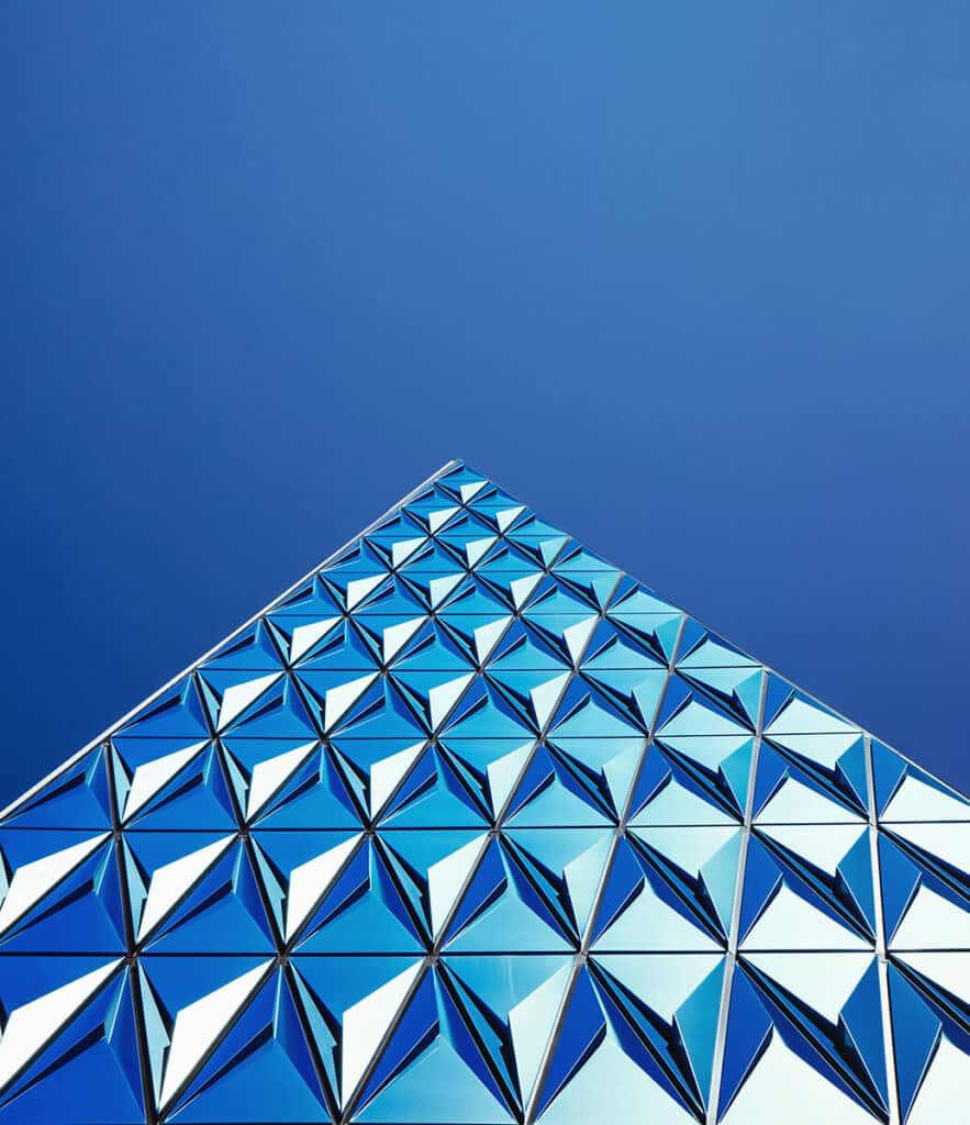 Upward view of a modern building facade with a geometric pattern of blue and white triangular panels against a clear blue sky.