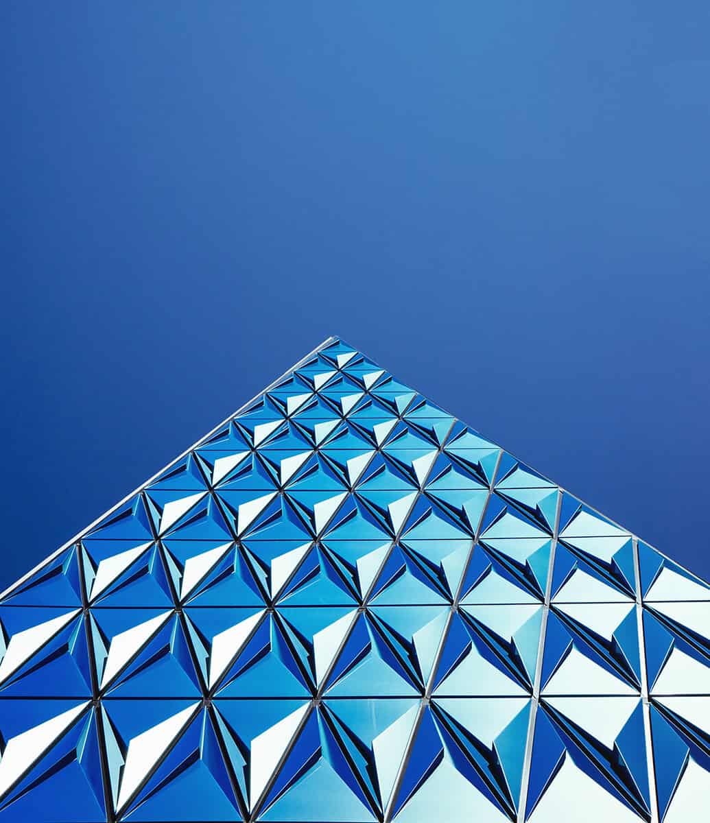 Upward view of a modern building facade with a geometric pattern of blue and white triangular panels against a clear blue sky.