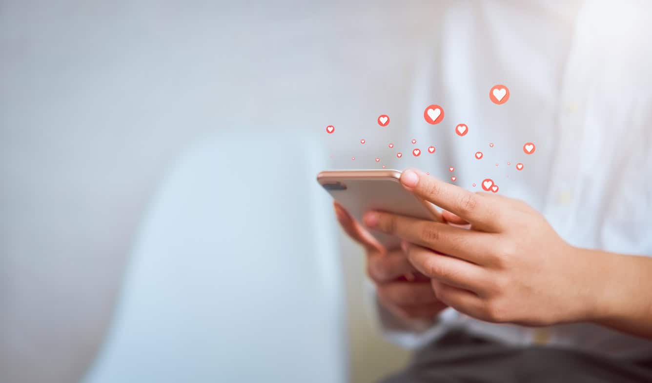 Close-up of hands holding a smartphone with red heart emojis floating upwards from the screen.
