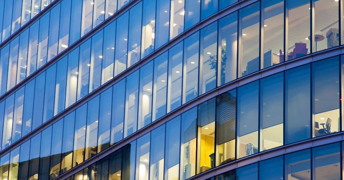 Exterior view of a modern glass office building with many interior lights on, reflecting a cool blue tone.