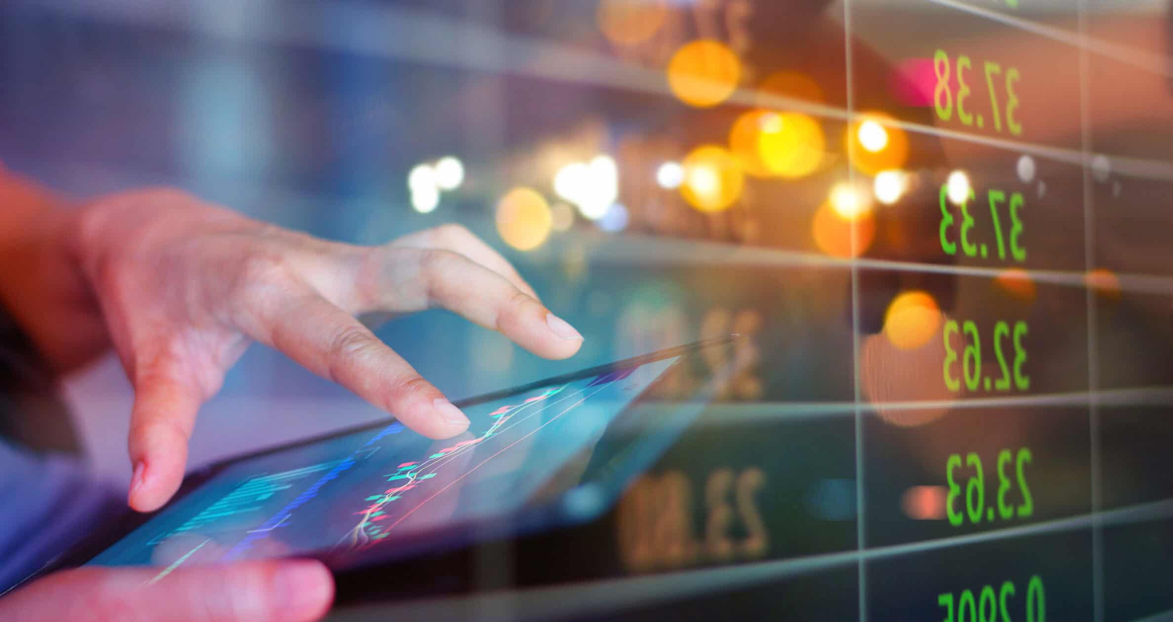 A hand touches a tablet screen displaying financial charts and graphs, with blurred lights and reflected numbers in the background.