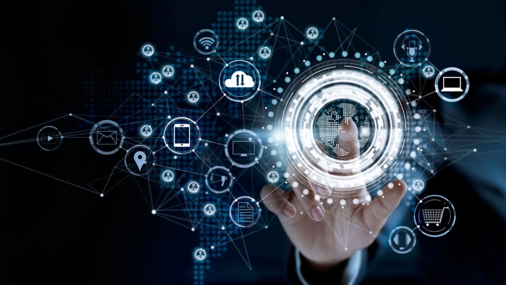 A hand interacting with a glowing digital interface featuring a globe and interconnected technology icons like cloud, Wi-Fi, smartphone, and shopping cart, against a dark background with a world map.