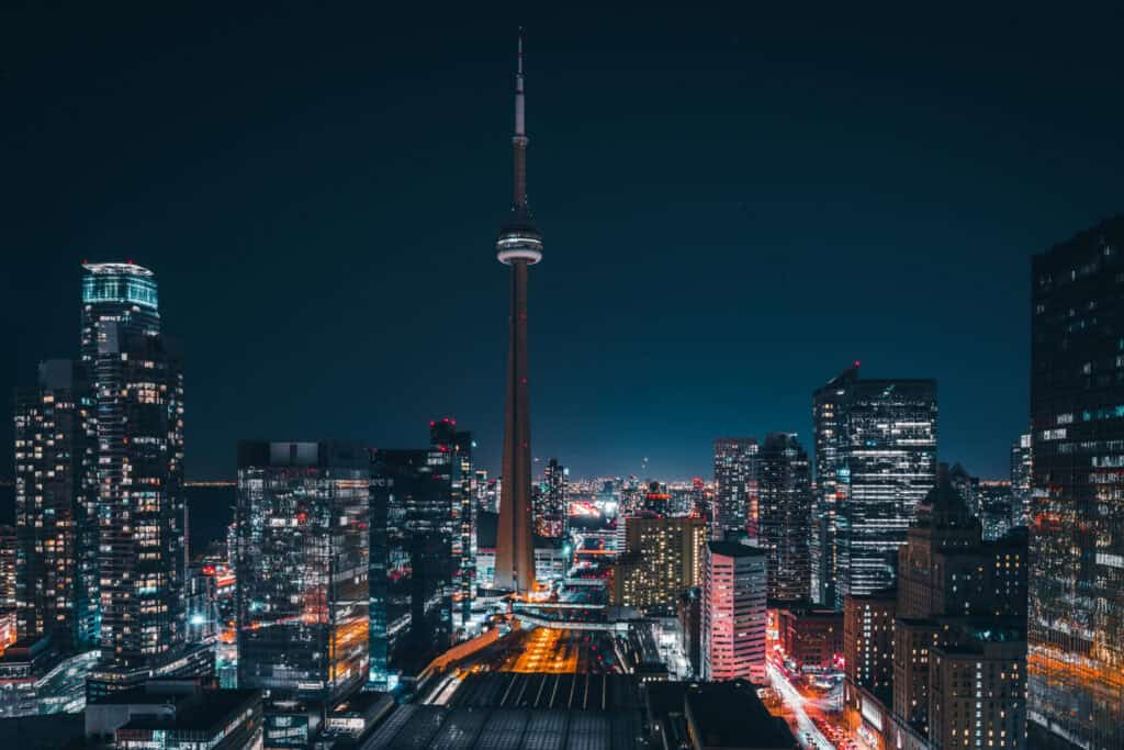 A panoramic night view of the Toronto skyline, featuring the brightly lit CN Tower surrounded by numerous illuminated skyscrapers and city lights.