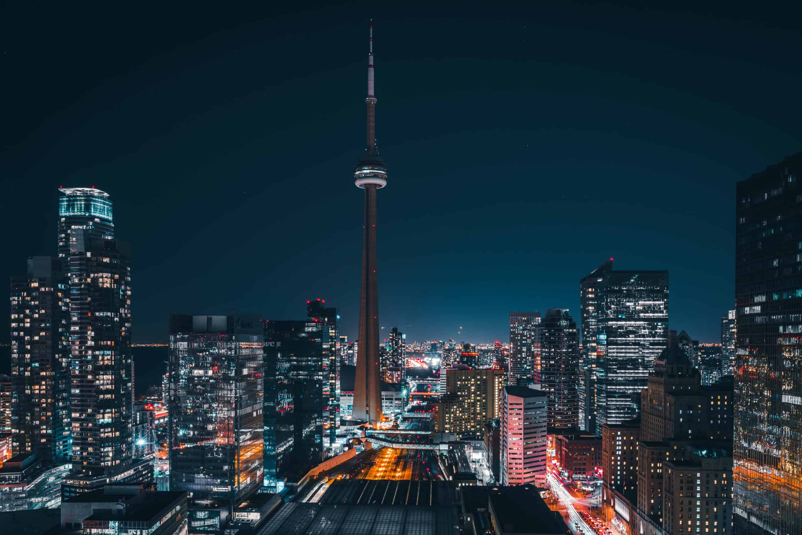 A panoramic night view of the Toronto skyline, featuring the brightly lit CN Tower surrounded by numerous illuminated skyscrapers and city lights.