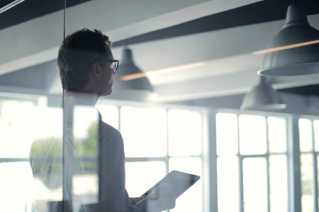 A man in glasses holds a tablet, looking out a bright window in a modern office.