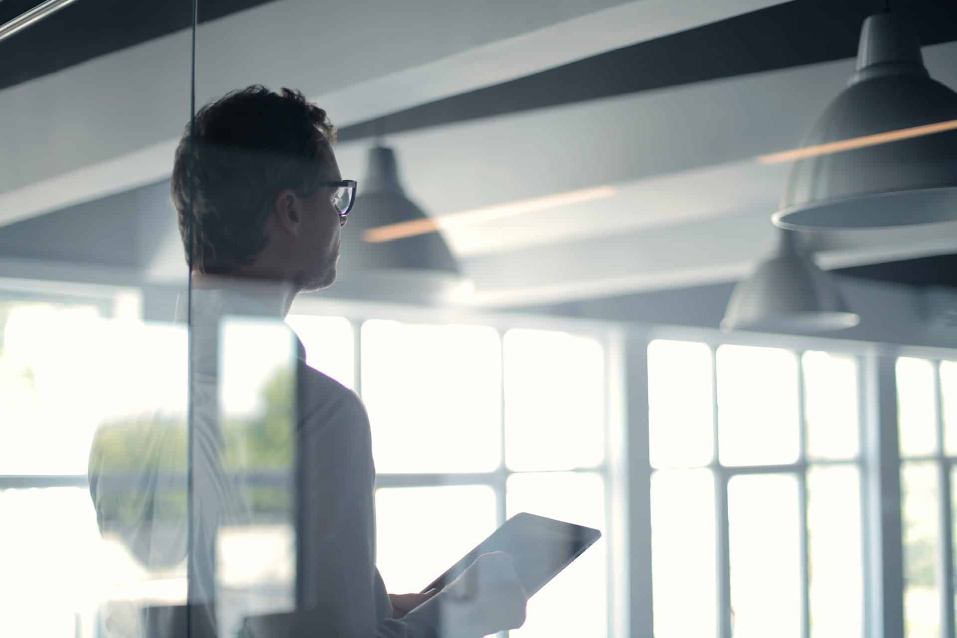 A man in glasses holds a tablet, looking out a bright window in a modern office.