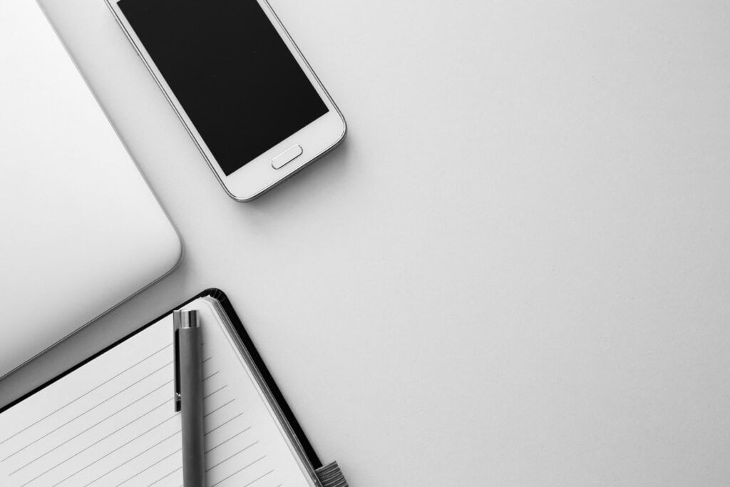 Overhead view of a silver laptop, white smartphone, open lined notebook, and silver pen on a light gray surface.