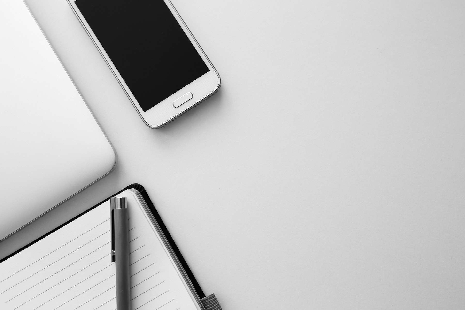 Overhead view of a silver laptop, white smartphone, open lined notebook, and silver pen on a light gray surface.