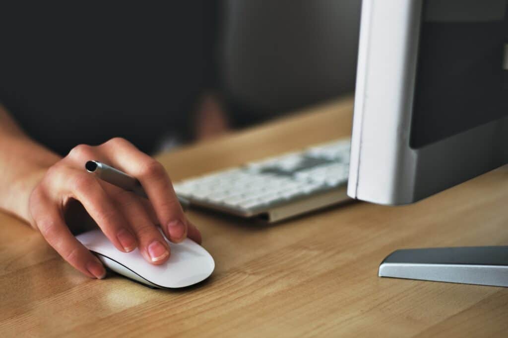 A person's hand holds a silver pen while using a white computer mouse on a wooden desk. A keyboard and monitor are blurred in the background.