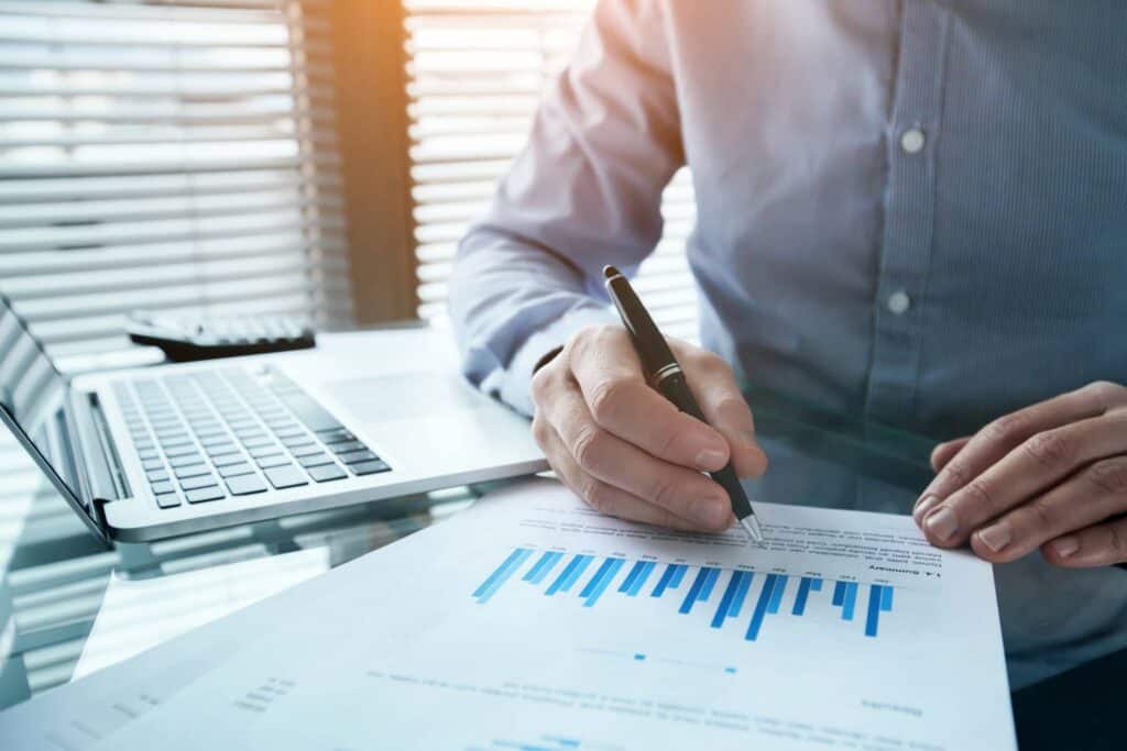 A person's hands holding a pen over a document with a bar chart, next to a laptop and calculator on a glass desk, with sunlit blinds in the background.