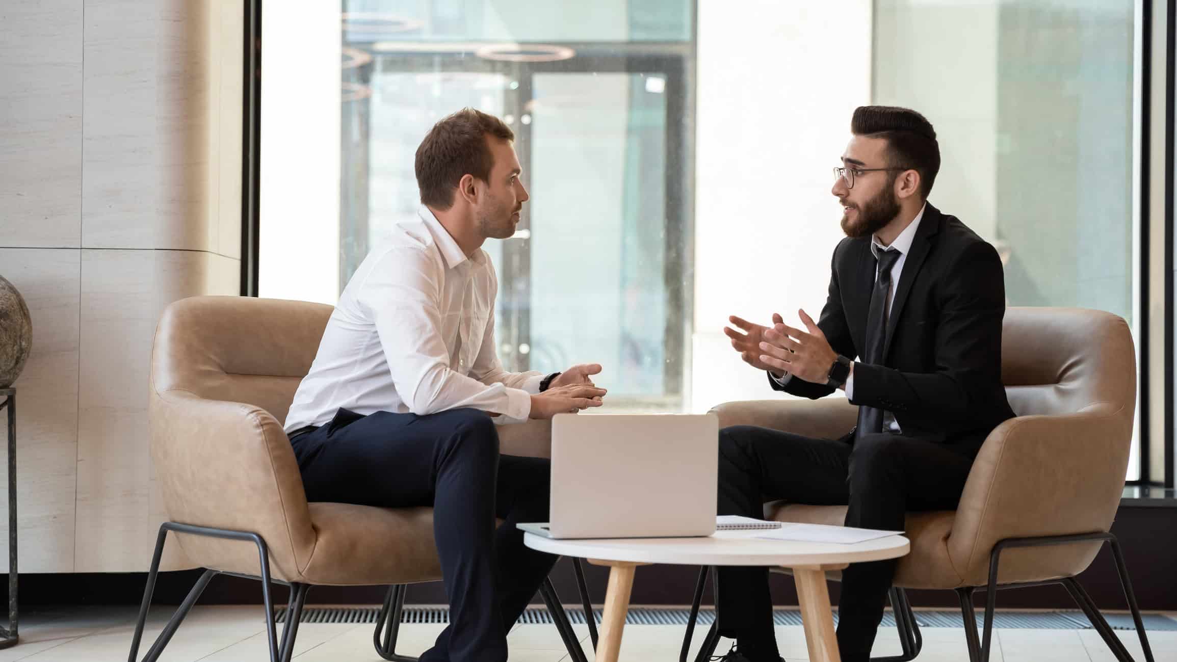 Two men in business attire sit in armchairs, facing each other and conversing, with a laptop on a table between them.