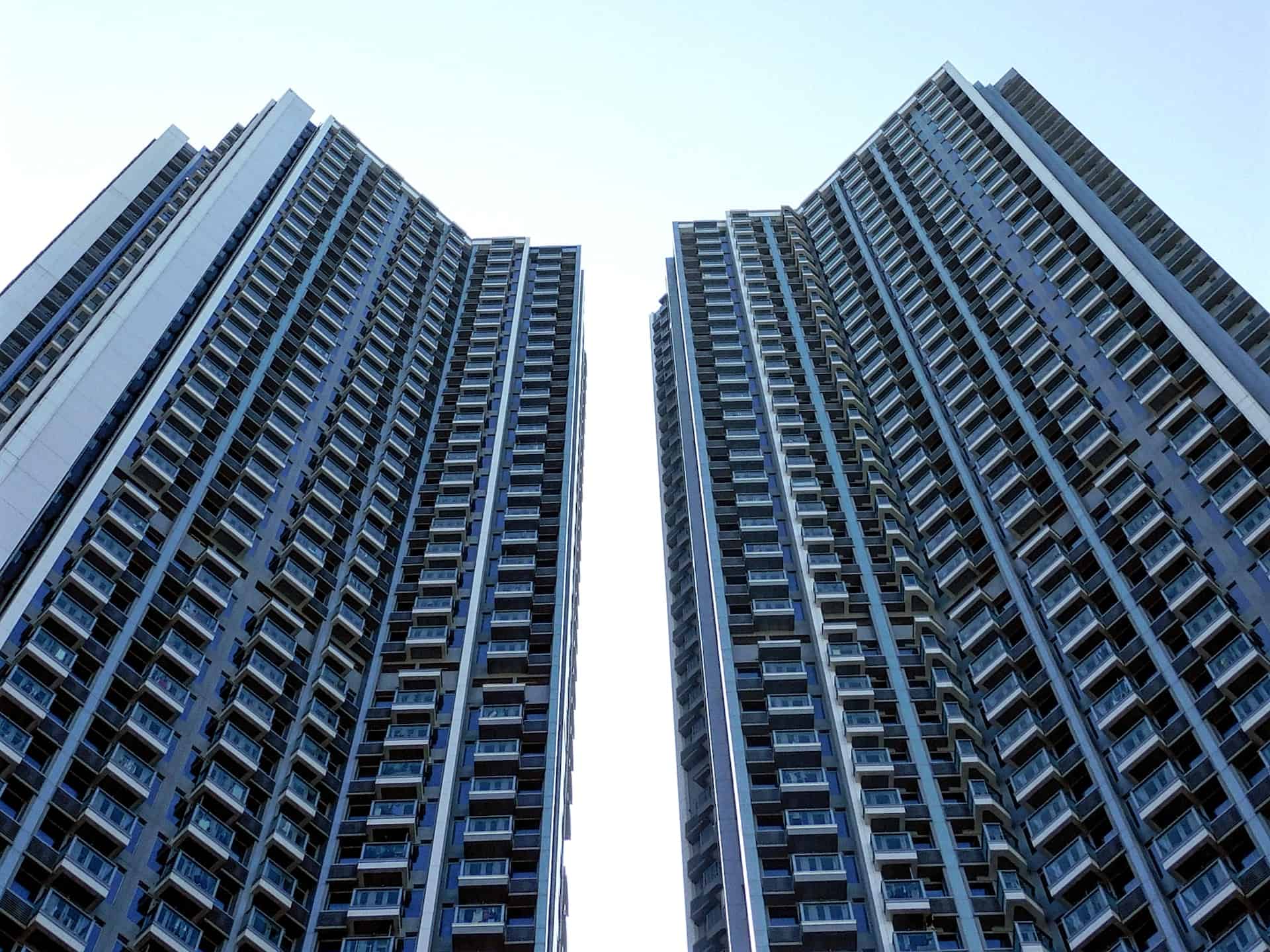 Two tall, grey and blue high-rise residential buildings seen from a low angle, with a bright sky between them.