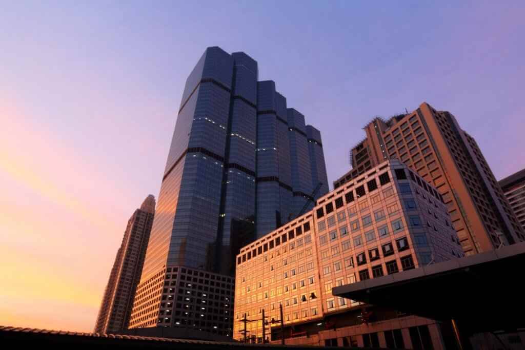 Low-angle view of multiple skyscrapers against a sky transitioning from orange to purple, with a large dark glass building prominent.