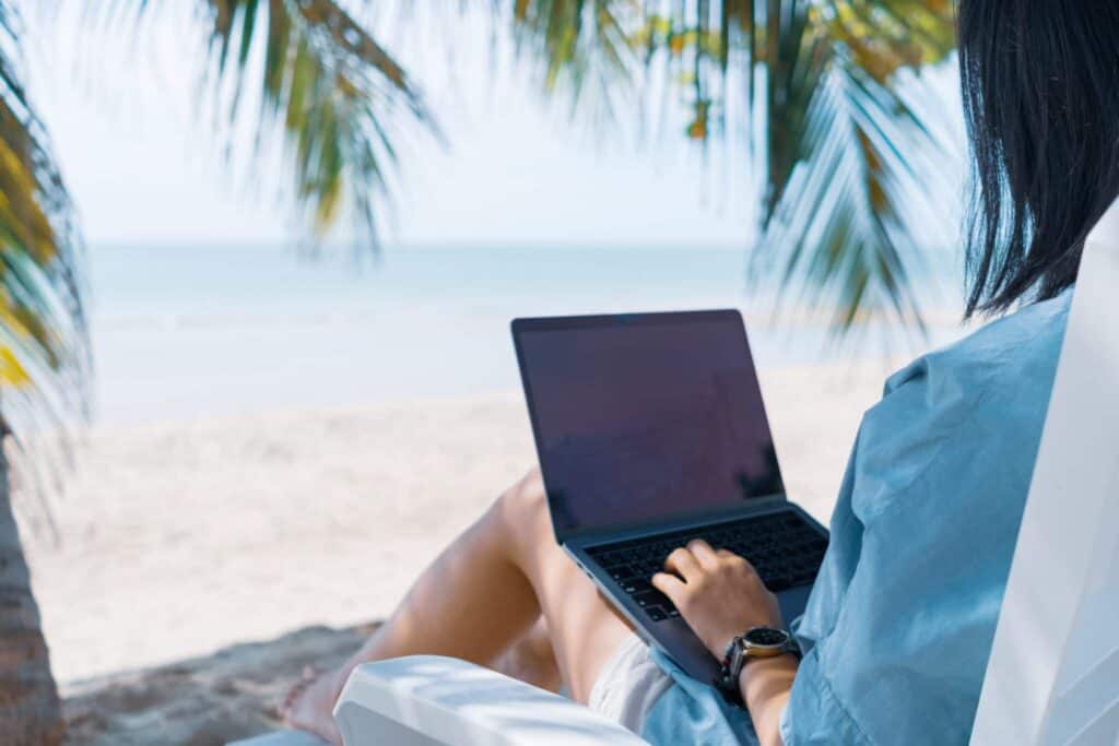 A person sits on a white beach chair, working on a laptop with the ocean and palm trees in the background.