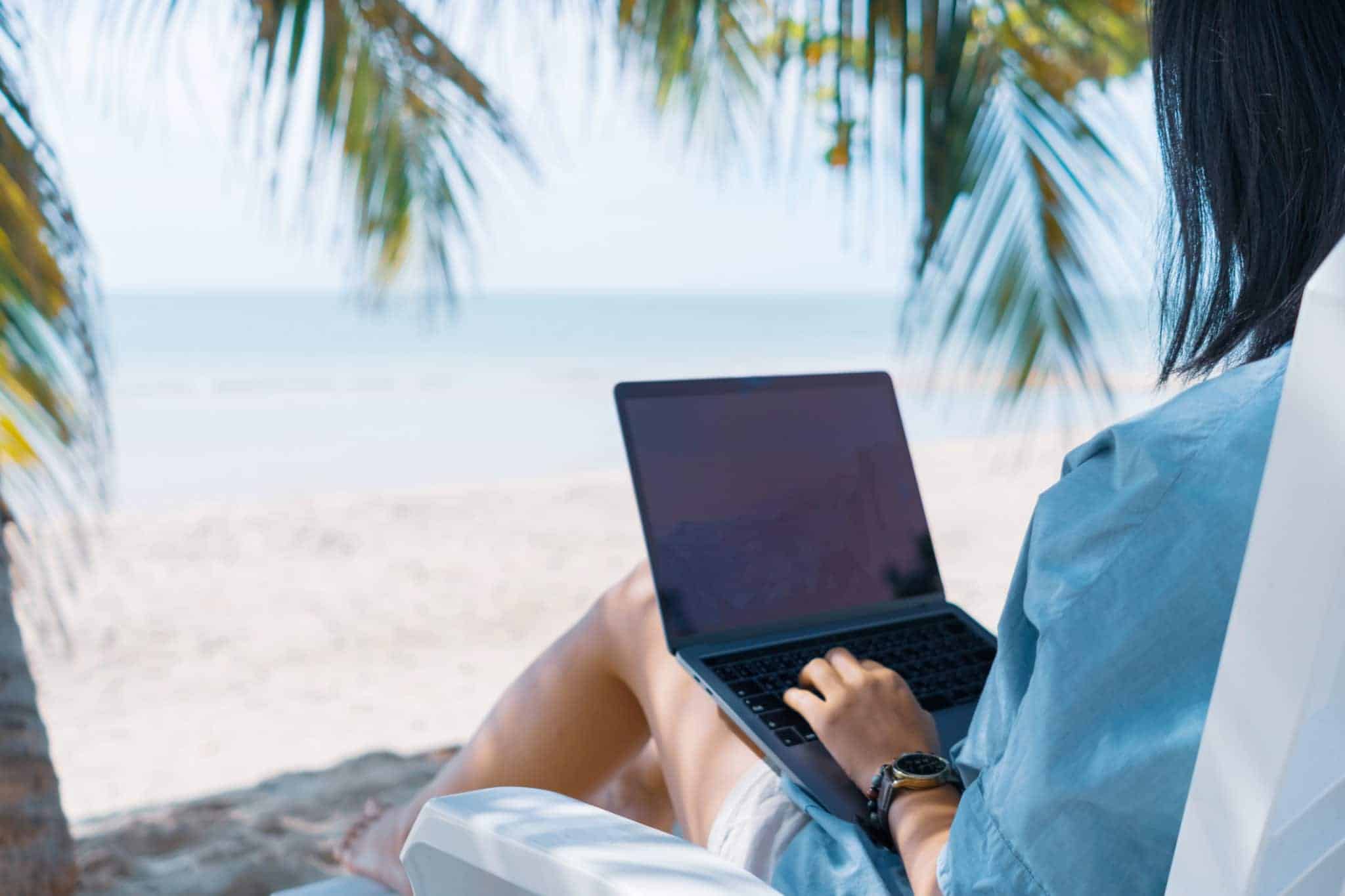 A person sits on a white beach chair, working on a laptop with the ocean and palm trees in the background.