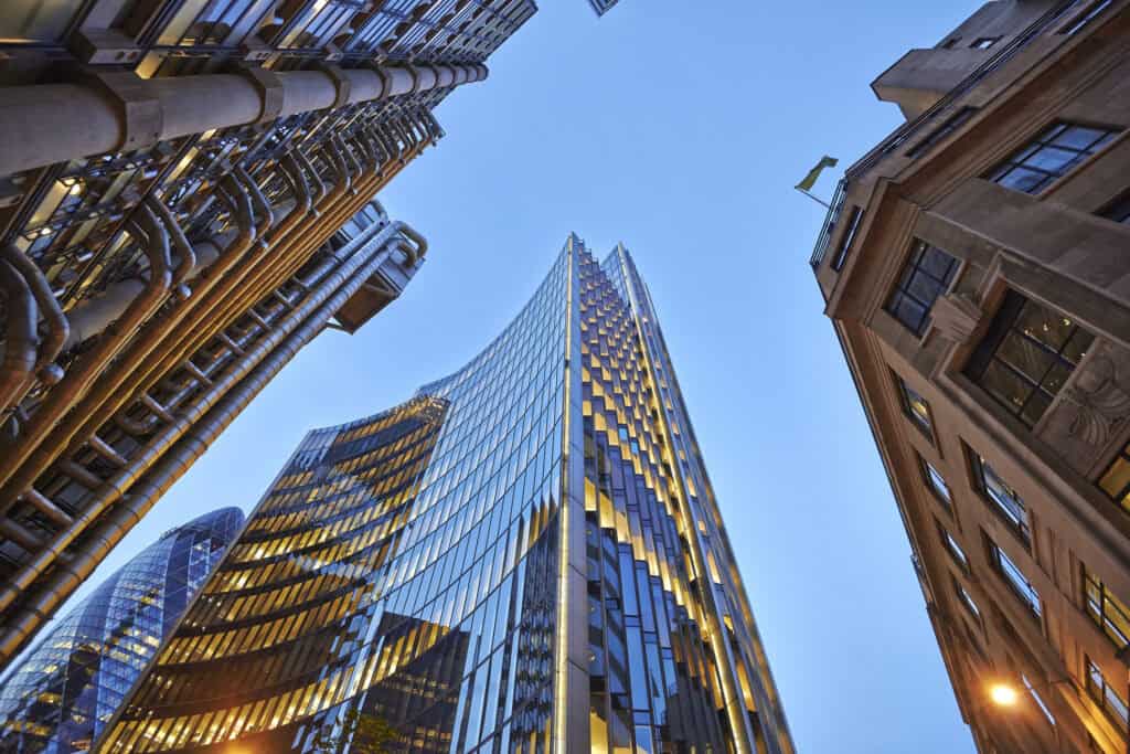 Low-angle view of multiple skyscrapers at dusk, including the Lloyd's Building, The Gherkin, and a modern glass tower, all illuminated against a deep blue sky.