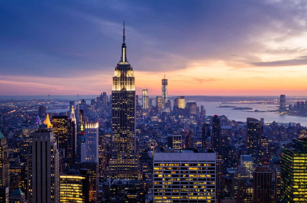 An aerial view of the New York City skyline at dusk, featuring the brightly lit Empire State Building and numerous other skyscrapers under a sky transitioning from deep blue to orange and pink.