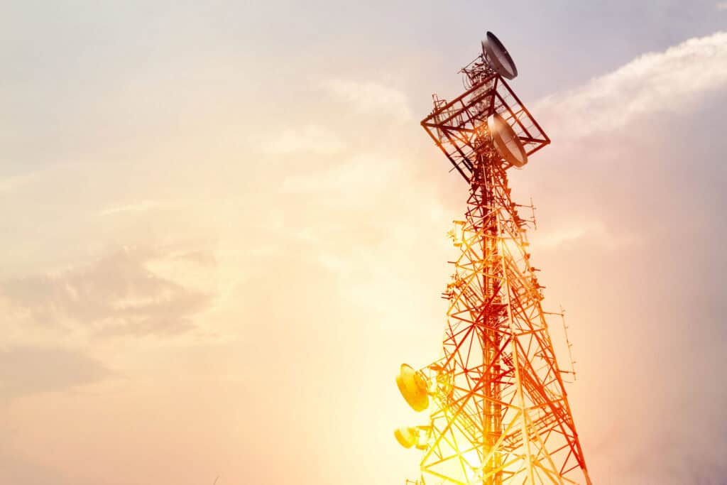 A tall communication tower with multiple antennas silhouetted against a bright, golden-orange sky at sunrise or sunset.