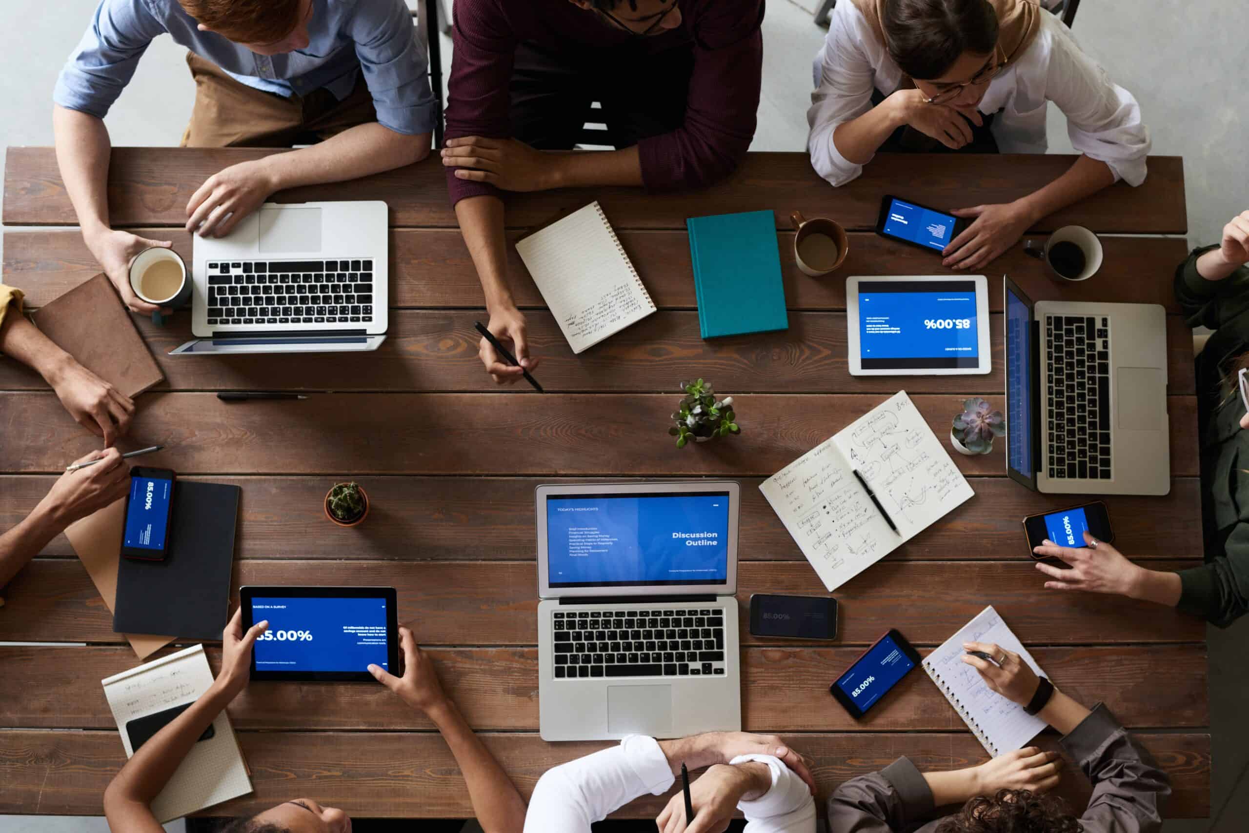 Overhead view of eight people working around a wooden table with laptops, tablets, notebooks, and smartphones, some displaying "85.00%" and "Discussion Outline".