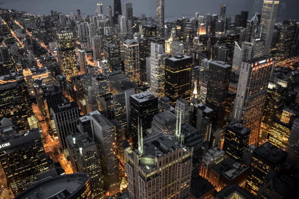 Aerial view of a city at night, showing numerous illuminated skyscrapers and streets with bright lights under a dark sky.
