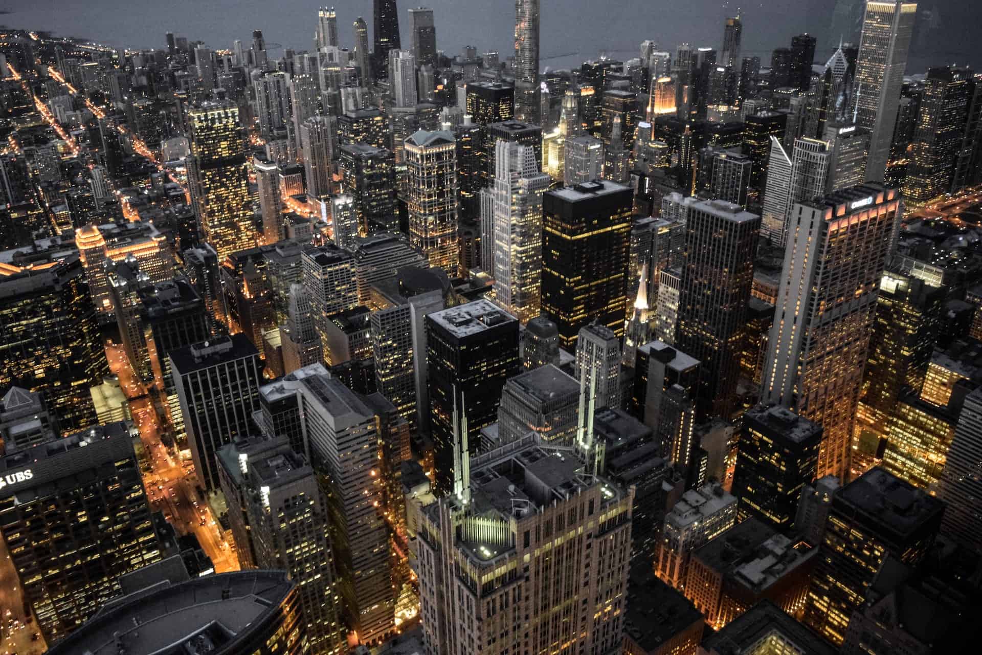 Aerial view of a city at night, showing numerous illuminated skyscrapers and streets with bright lights under a dark sky.