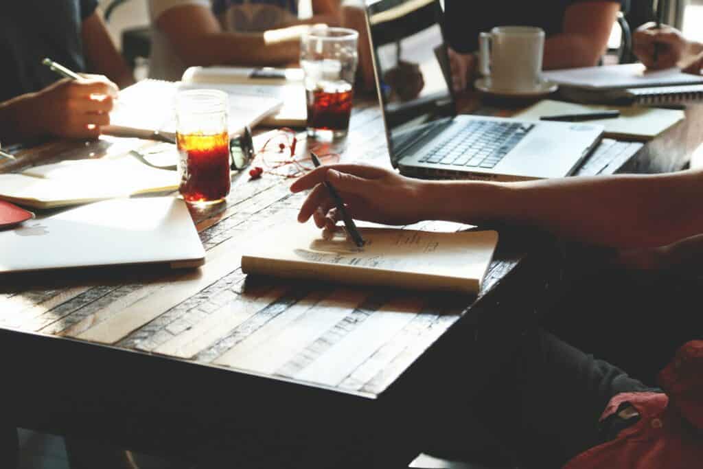 People working at a wooden table with laptops, notebooks, pens, and drinks.