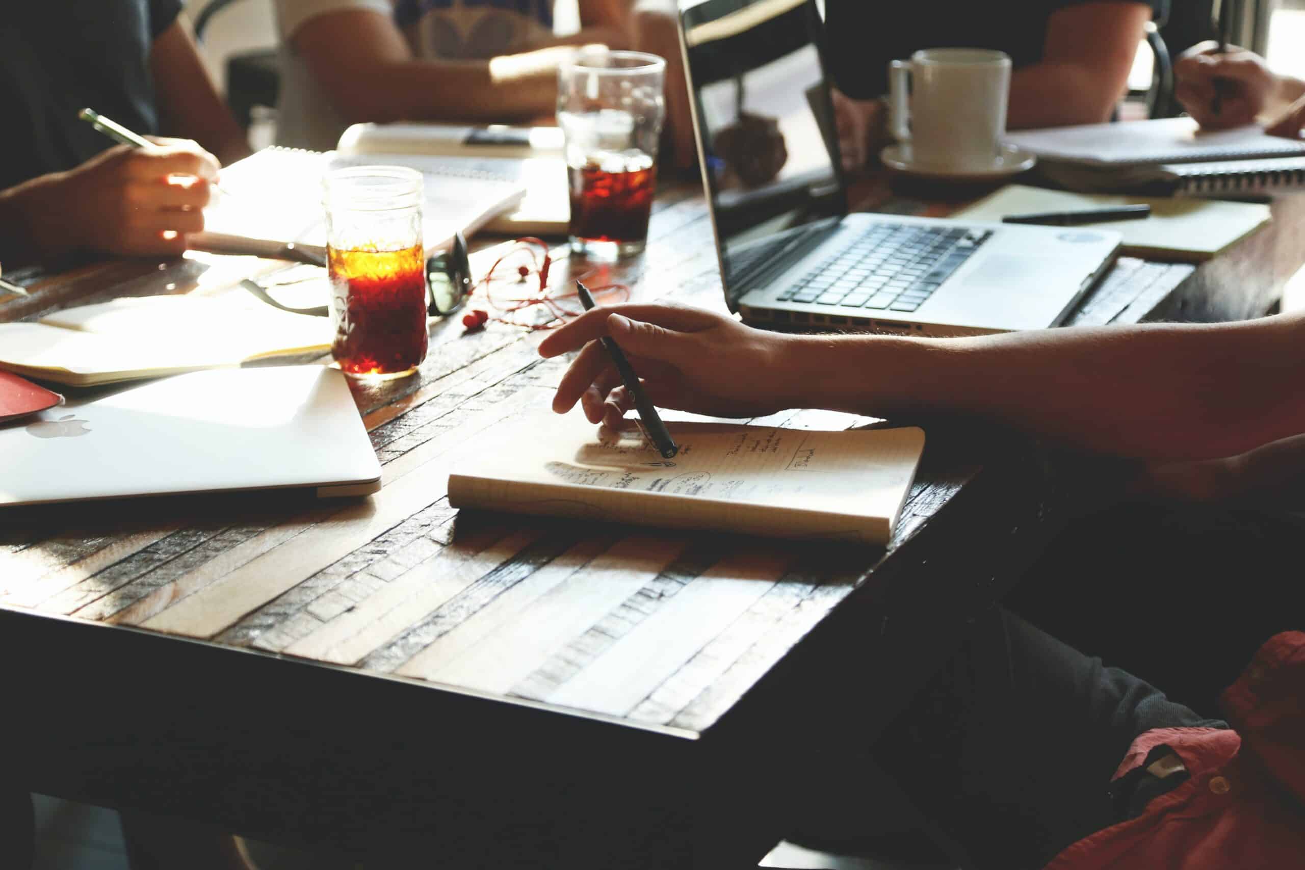 People working at a wooden table with laptops, notebooks, pens, and drinks.