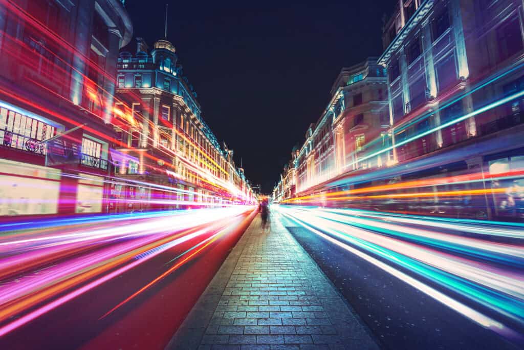 Long exposure photo of a city street at night with colorful light trails from vehicles and illuminated buildings.