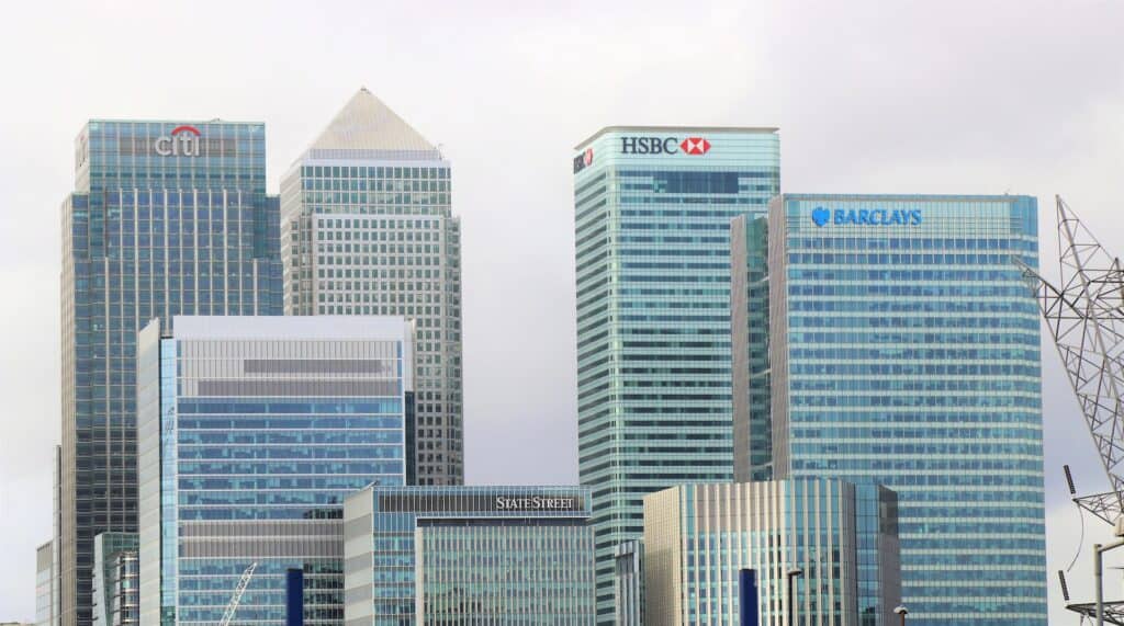 A cluster of glass skyscrapers under an overcast sky, featuring the logos of Citi, HSBC, Barclays, and State Street.