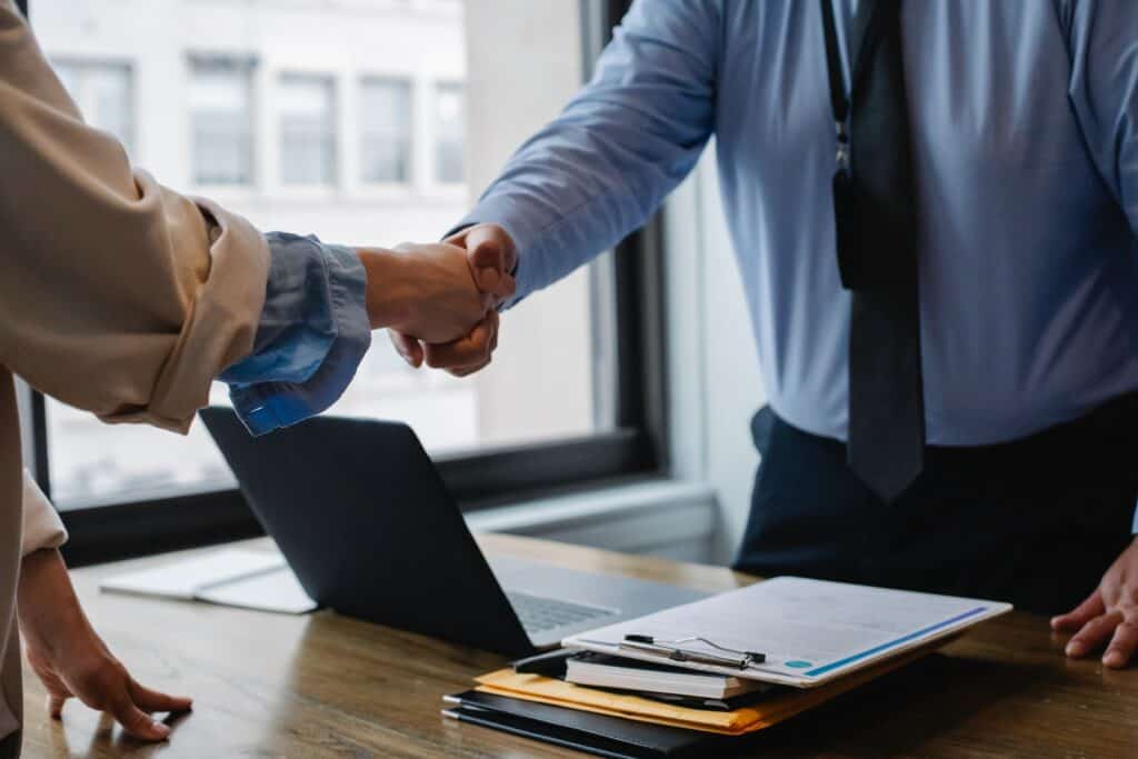 Two people shaking hands over a wooden desk with a laptop and documents, in front of a window.