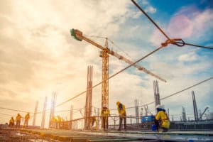 Construction site with workers in hard hats and vests, a large yellow tower crane, and rebar structures under a bright, cloudy sky.