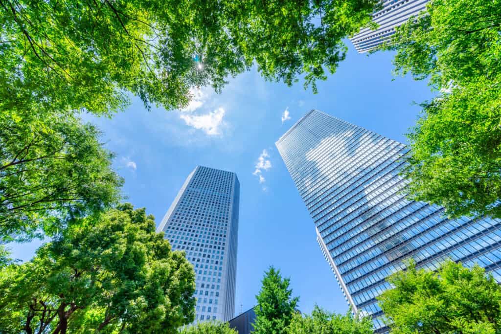 Two tall, modern skyscrapers with reflective glass facades, seen from a low angle against a bright blue sky with scattered clouds, framed by lush green tree leaves.